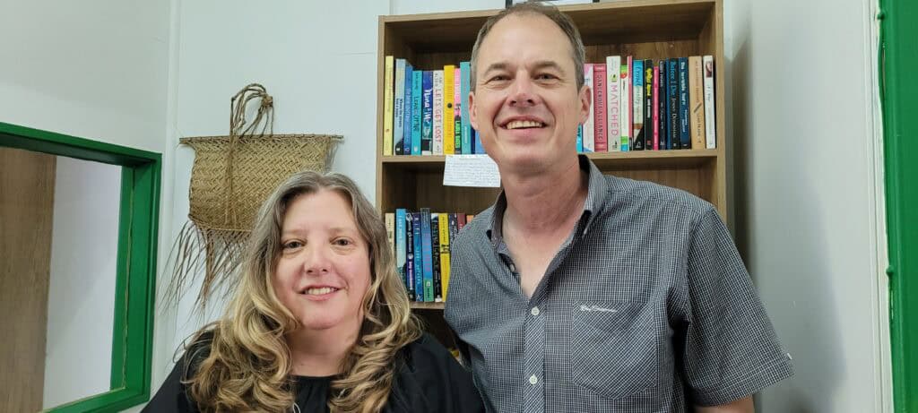 Two people standing before a bookshelf, one woman with blond hair and a tall man.