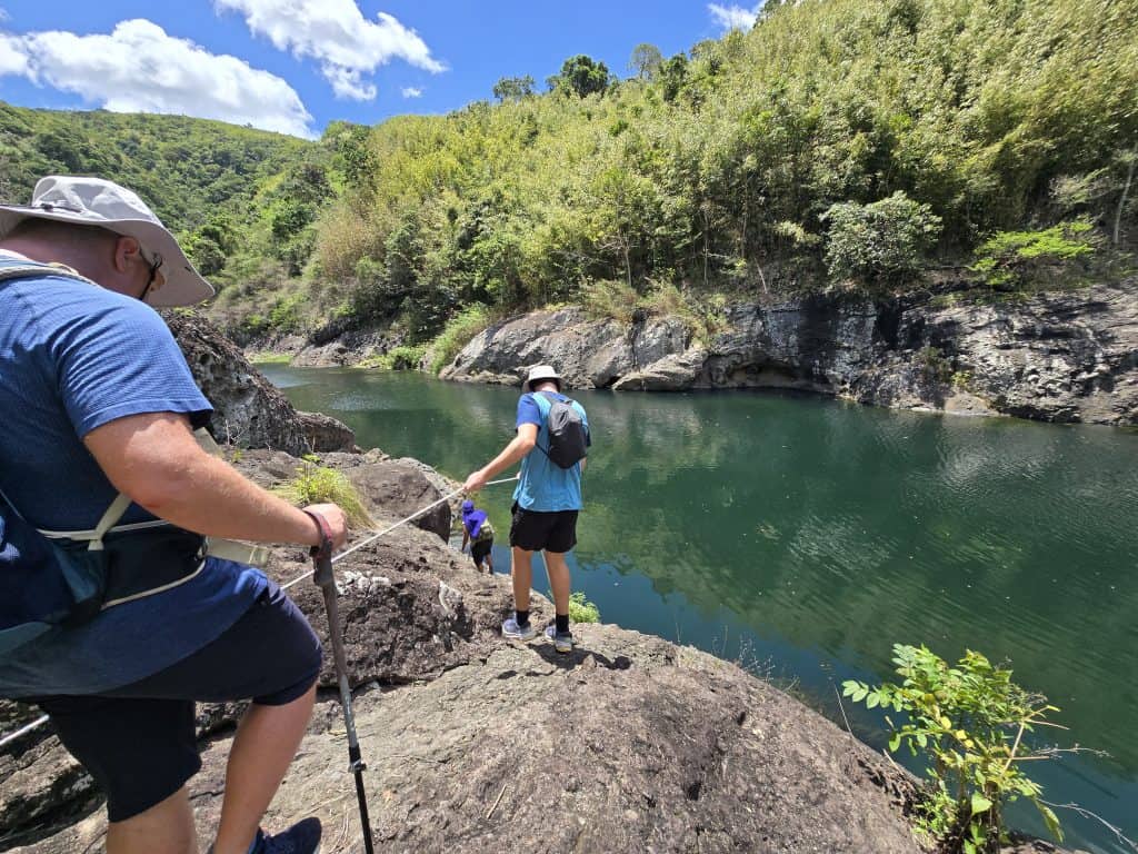 Trekkers hang on to rope as they walk over rocks near a river